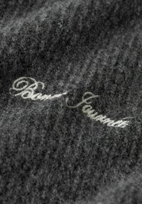 Grått strukturerat stickat ull med en mjuk, fluffig yta. Har vit broderad text "Bari Javan" i en elegant kursiv stil.