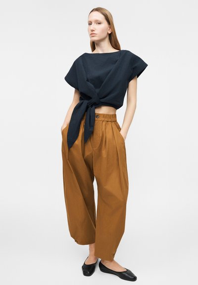 Γυναίκα που στέκεται με τα χέρια στις τσέπες, φοράει άνετο καφέ παντελόνι με ψηλή μέση, μαύρο crop top με κορδόνια και μαύρα επίπεδα παπούτσια, μπροστά σε απλό φόντο.