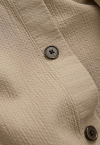 Beiges Gewebe mit strukturiertem Muster, versehen mit einem dunkelbraunen Knopf mit vier Löchern, der auf einem Hemd oder Kleidungsstück positioniert ist.