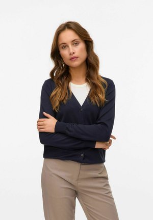 Cardigan bleu marine avec un col en V, fermeture à un bouton, poignets côtelés et une texture douce, associé à un pantalon beige clair.