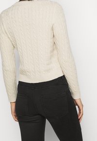Gestrickter cremefarbener Pullover mit einem strukturierten Zopfmuster, langen Ärmeln und geripptem Saum, kombiniert mit schwarzen, eng anliegenden Jeans mit Gesäßtaschen.