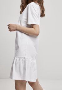 Žena v krátkých rukávech bílých šatech s nařaseným volánem na spodním okraji, stojící bokem proti jednoduchému světlému šedému pozadí.