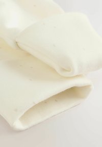 Kremno obarvani pleteni material z gladko teksturo in majhnimi svetlo bež pikicami. Manšetni robovi so zavihani in robovi mehki.