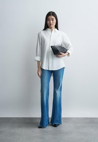 Γυναίκα με μακριά σκούρα μαλλιά φοράει λευκό πουκάμισο με κουμπιά και μπλε τζιν καμπάνα, κρατώντας μια μαύρη τσάντα φάκελο πάνω σε απλό γκρι φόντο.