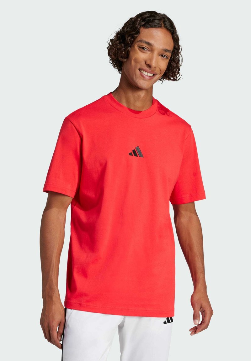 T-shirt in cotone rosso con maniche corte, caratterizzata da un logo Adidas nero sul petto, scollo rotondo e vestibilità standard.