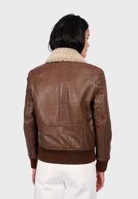 Chaqueta bomber de cuero marrón con cuello de borrego, puños y dobladillo acanalados; textura suave con costuras detalladas, utilizada sobre pantalones blancos.