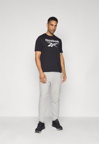 T-shirt noire Reebok avec logo blanc, pantalon de survêtement gris et baskets noires. Le modèle se tient contre un fond neutre dans une pose détendue.