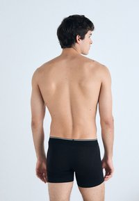 Black boxers made from a soft fabric, featuring a white elastic waistband. The model is viewed from the back, with a natural skin tone.