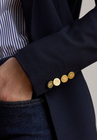 Hånd i jeanslomme, iført navy blazer med fire gyldne knapper med logo på ærmet og en blå- og hvidstribet skjorte under.