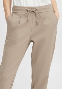 Beige Jogger-Hosen aus weichem Material, mit einem Kordelzugbund, seitlichen Taschen und dezenten Falten für zusätzliche Textur.