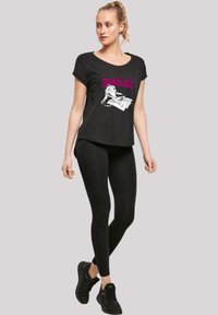 Svart grafisk t-skjorte med avslappet passform, med en hvit illustrasjon og rosa tekst som sier "Jeg gjør ikke morgener." Kombinert med svarte leggings og sneakers.