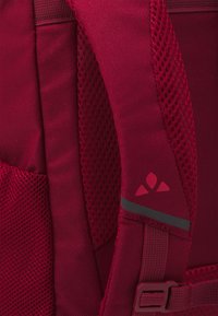 Bordo ruksak s teksturiranom mrežastom trakom, glatkim materijalom i malim logotipom. Uključuje podesivu opremu za prilagodbu.