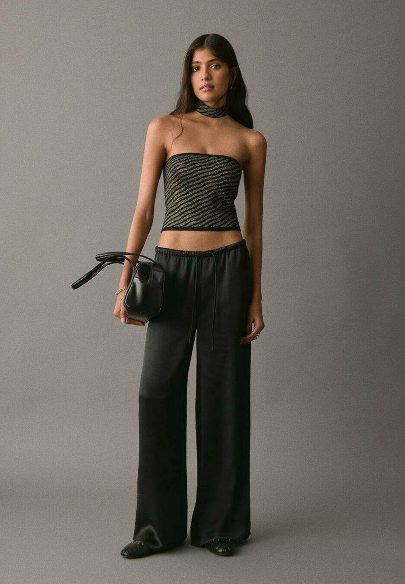 Čierny top bez ramienok s metalickými pruhmi v kombinácii s voľnými čiernymi nohavicami, doplnený malou čiernou kabelkou a kruhovými náušnicami.
