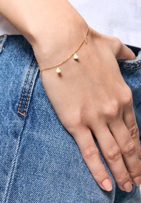 Złota bransoletka z trzema małymi sercami, noszona na nadgarstku na tle dżinsu, ukazująca delikatny wzór i gładką teksturę.