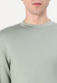 Camisa de manga longa verde-claro com decote canelado e textura suave, apresentando costuras mínimas para um design limpo.