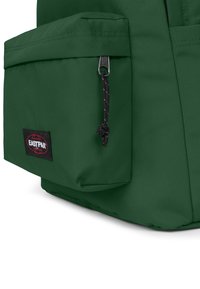 Grön Eastpak-ryggsäck med texturerad tyg, som har en framficka, svart dragkedja och ett märkesetikett. Enkelt design med funktionella detaljer.