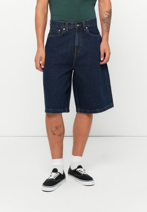 Dark blue denim shorts with a loose fit, featuring a button and zipper closure, five pockets, and rolled hems, worn with black sneakers.