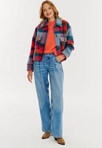 Pestrý károvaný sakó z vlnenej zmesi s dvoma prednými vreckami, oblečené cez oranžový sveter a v kombinácii s vysokými modrými džínami.