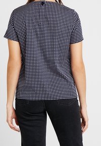 Tmavě modrý top s krátkým rukávem a bílým čtvercovým vzorem, s kulatým výstřihem a malým knoflíkovým zapínáním na zadní straně.