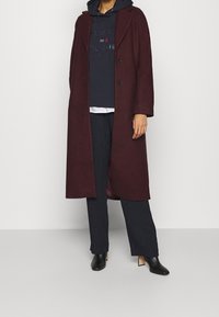 Bordowy długi płaszcz z klapowym kołnierzem, dwoma guzikami i otwartym przodem. Nosi się go na granatową bluzę z kapturem i ciemne spodnie, do tego czarne botki na obcasie.