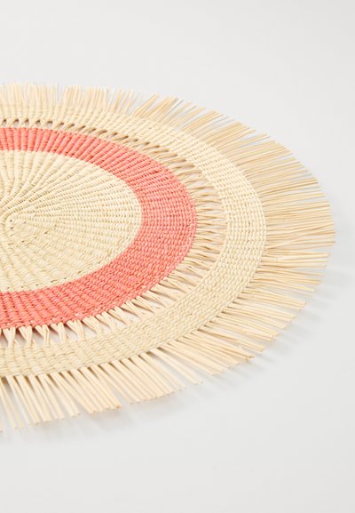 Round, woven placemat in natural fibers, featuring concentric circles in cream and coral, with fringe detailing around the perimeter.