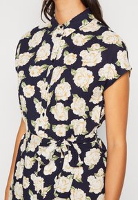 Marinblå blommig klänning med krämfärgade rosor, korta ärmar, krage och knytbart midjeband, tillverkad av ett lätt tyg.