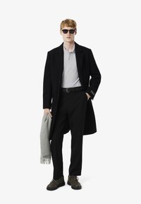 Svart kappa över en grå polo, svarta byxor och olivgröna sneakers. Håller en ljusgrå halsduk, bär mörka solglasögon. Slim fit-design.