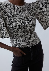 Hedvábná blůza s květinovým potiskem v krémové barvě s černými skvrnami, širokými rukávy a přiléhavým pasem, zkombinovaná s černými džíny s vysokým pasem.