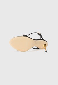 Black high-heeled sandal with a slender strap design and gold-tone hardware. Features a smooth beige sole and a pointed heel with a decorative accent.
