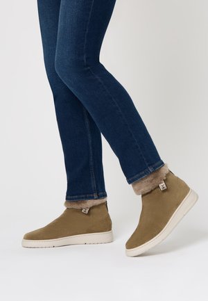 Hellbraune Wildleder-Stiefeletten mit weichem Futter aus Fell, weißen Gummisohlen und Branding-Detail an der Ferse. Getragen mit engen blauen Jeans.