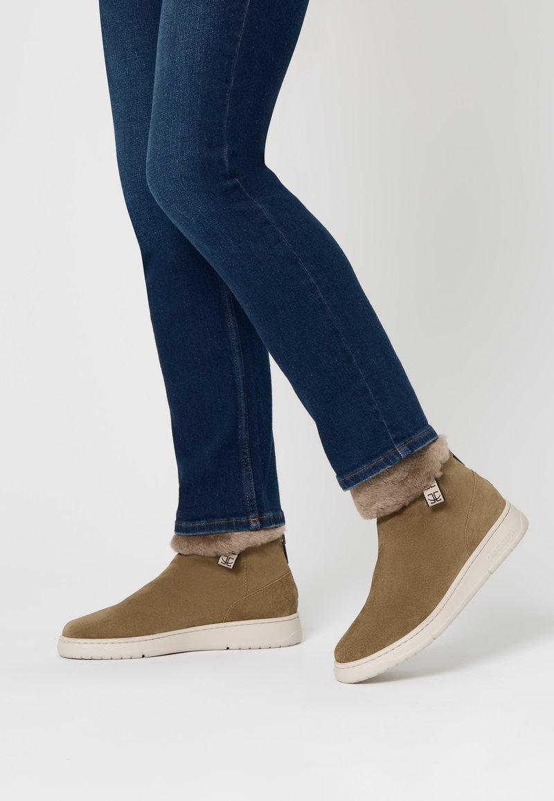 Stivaletti alla caviglia in suede color cammello con morbido rivestimento in pelliccia, suole in gomma bianca e dettaglio del marchio sul tallone. Indossati con jeans blu aderenti.