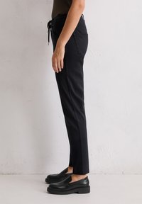 Zijaanzicht van een persoon in zwarte slim-fit broek met een strikband in de taille en zwarte leren instappers tegen een effen muur.