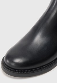 Stivale alla caviglia in pelle nera con punta rotonda, finitura testurizzata e dettagli di cucitura a contrasto lungo la suola. Aspetto liscio e lucido.