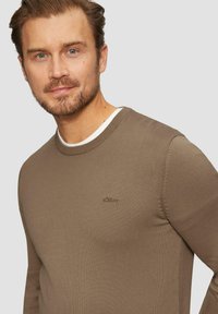 Rjav, rebrast pulover s krožnim izrezom z majhnim vezenim logotipom na prsih. Dolgi rokavi z oprijetim krojem. Obložen preko bele srajce.