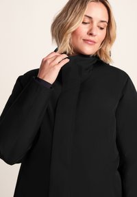 Woman adjusting the collar of a black high-neck jacket, looking down with a neutral expression against a plain light background.