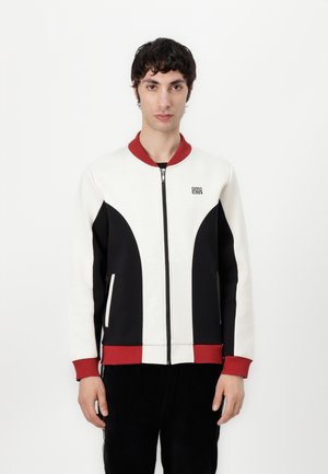 Hvit og svart zip-up jakke med røde detaljer, med stående krage, sidelommer og en logo på brystet. Glatt stoffkvalitet.