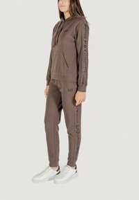 Mujer joven con sudadera EA7 Emporio Armani marrón y joggers a juego, usando zapatillas blancas, de pie frente a un fondo liso.