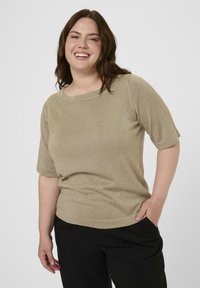 Glunderende vrouw met bruin golvend haar draagt een gouden top met korte mouwen en zwarte broek, staat met één hand in haar zak tegen een effen achtergrond.