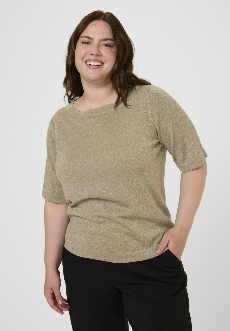 Mujer sonriente con cabello castaño y ondulado que lleva una blusa dorada de manga corta y pantalones negros, de pie con una mano en el bolsillo frente a un fondo liso.