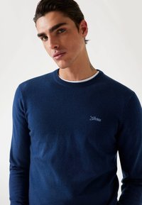 Sötétkék, crewneck pulóver, puha anyagból készült, hosszú ujjal és testhez álló szabással. A mellkasán hímzett "Guess" logóval rendelkezik.