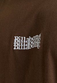Hnedé bavlnené tričko s bielym vyšitým textom, ktorý znie "Billabong" v opakovanom, zrkadlovom dizajne po ľavej strane.