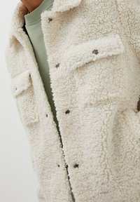 Kræmfarvet fleecejakke med en blød tekstur, der har lappelommer og sorte knapdetaljer. Båret over en grøn sweater.