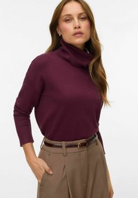 Maglione a collo alto bordeaux con maniche lunghe, abbinato a pantaloni beige a vita alta e una cintura marrone. Tessuto morbido e liscio.