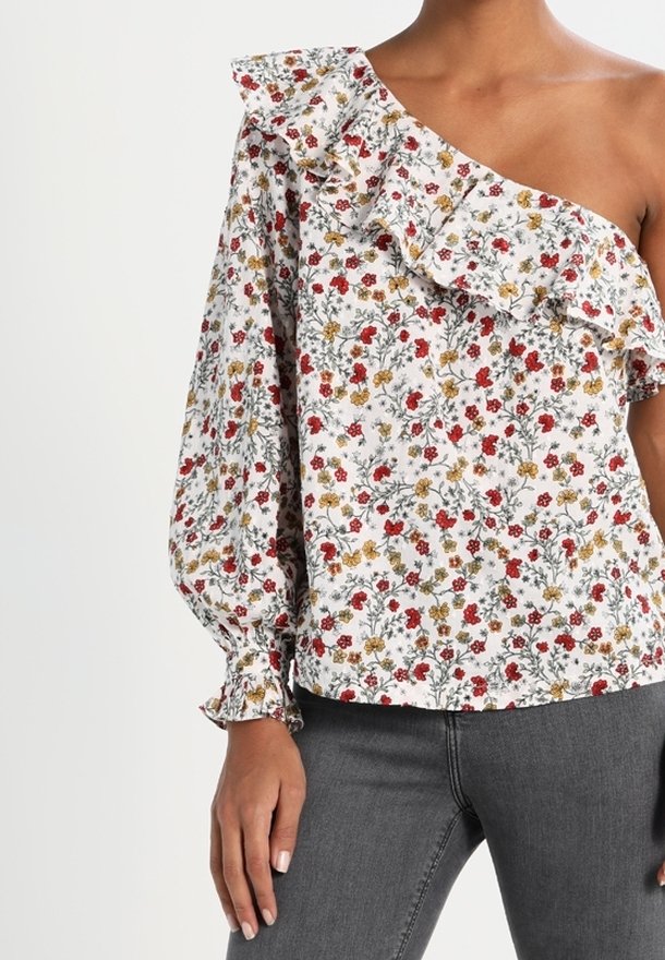 Femme portant un chemisier blanc asymétrique avec imprimé floral rouge et jaune et détails froncés, associé à un jean gris.