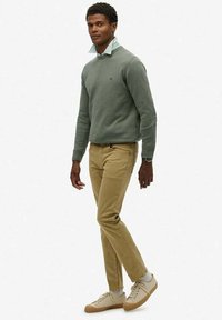 Suéter de punto verde sobre una camisa de cuello azul claro y blanca a rayas, combinado con chinos beige y zapatillas claras, de pie frente a un fondo liso.