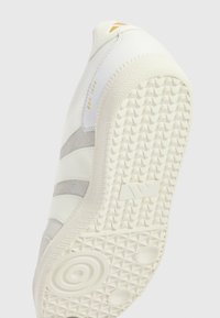 Sneaker blanc avec des accents en suède et cuir gris. Semelle en caoutchouc avec un motif texturé. Présente un logo et "EST. 1994" en doré.