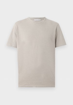 Άνδρας με μπεζ μπλουζάκι, ανοιχτό κρεμ σορτς, λευκές κάλτσες και κρεμ αθλητικά παπούτσια, στέκεται με τα χέρια στις τσέπες κοιτάζοντας αριστερά.