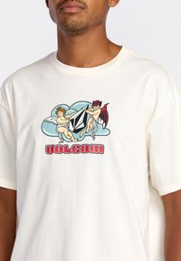 Camiseta de algodón color crema con un gráfico de ángeles y demonios sosteniendo un logotipo negro, con el texto en rojo "VOLCOM" en negrita debajo.