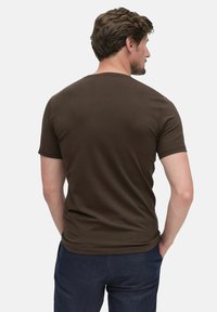 Homme avec des cheveux bruns courts portant un t-shirt uni marron à manches courtes et un pantalon bleu foncé, vu de dos sur fond blanc.