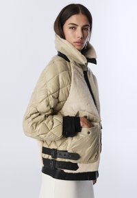 Femme en veste matelassée beige avec des empiècements en fourrure et des sangles en cuir noir, debout de profil, main dans la poche, sur un fond clair.